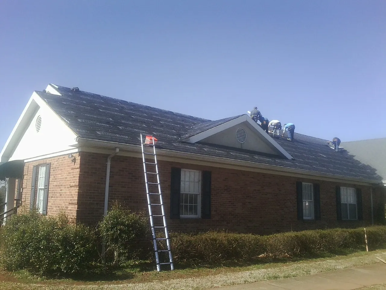 Crew tearing shingles off a brick ranch roof with a ladder set — Metal Roof Repair work by our crew in Biggs